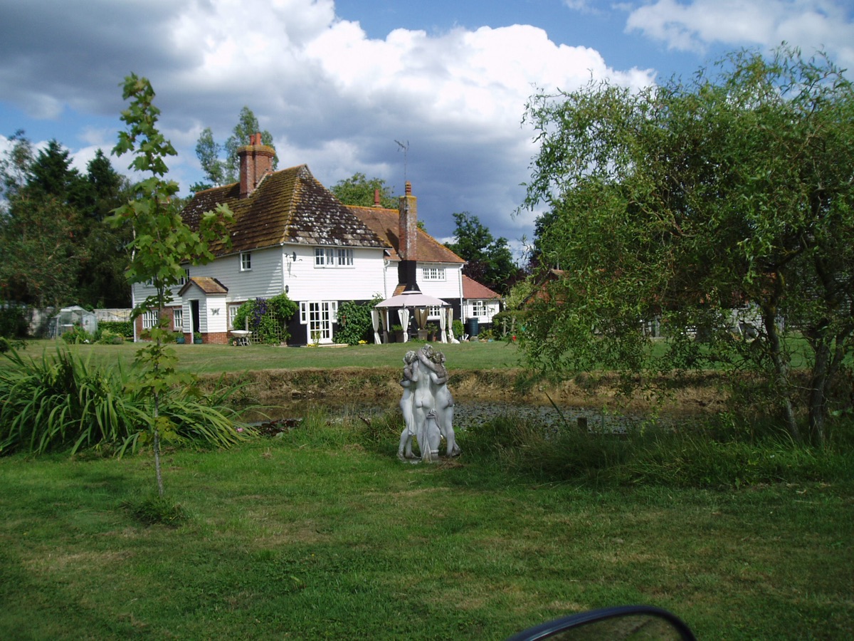 The weatherboarded farmhouse at Goffsland Farm seen from across the garden and pond, with a willow to the right and cloud building overhead